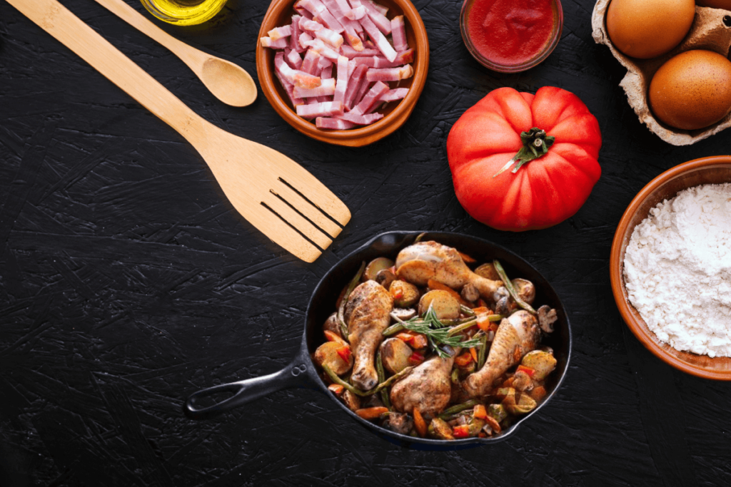 "Pros & cons of using cast iron pots and pans ,Overhead view of a cast iron pan filled with seasoned chicken drumsticks and vegetables, surrounded by wooden cooking utensils, a fresh tomato, flour, eggs, sliced bacon, and tomato sauce on a dark textured surface."