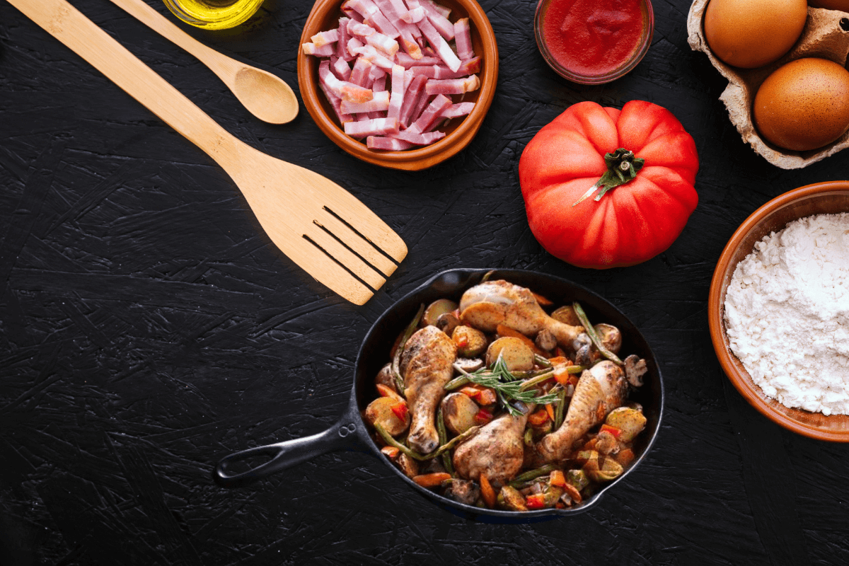 "Overhead view of a cast iron pan filled with seasoned chicken drumsticks and vegetables, surrounded by wooden cooking utensils, a fresh tomato, flour, eggs, sliced bacon, and tomato sauce on a dark textured surface."